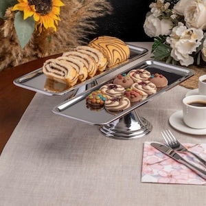 May include: A two-tiered, silver-colored serving tray holds an assortment of pastries and cookies. The top tier displays sliced rolls and a swirl pastry. The bottom tier features decorated cookies. A cup of coffee and silverware are also visible.