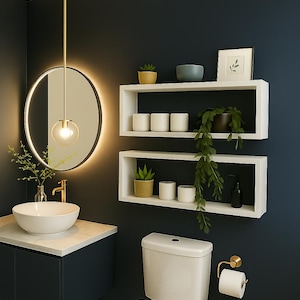 Op de afbeelding: Modern badkamerontwerp met een ronde spiegel met achtergrondverlichting, een witte wastafel en zwevende planken met planten en decoratieve items. Het kleurenschema omvat marineblauwe muren, witte armaturen en gouden accenten.