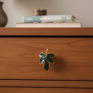 May include: A green and gold maple leaf cabinet knob fixed to a wooden drawer. The leaf-shaped handle adds a touch of nature-inspired design to the furniture. The drawer is made of wood.