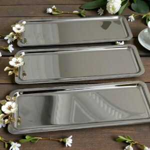 May include: Three rectangular stainless steel serving trays. Each tray has a floral accent with white flowers and gold-colored leaves on one corner. The trays are arranged on a wooden surface.