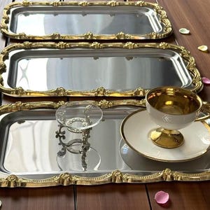 May include: Three rectangular silver trays with gold-colored ornate edges. One tray holds a teacup and saucer with gold accents, and a small glass stand. The trays are arranged on a wooden surface.