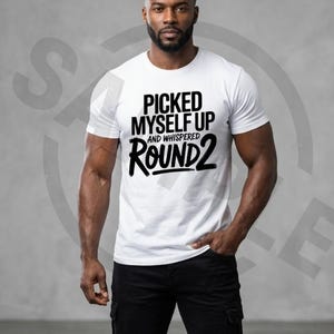 May include: A white t-shirt with the text "PICKED MYSELF UP AND WHISPERED ROUND 2" in black. The person is wearing black trousers.