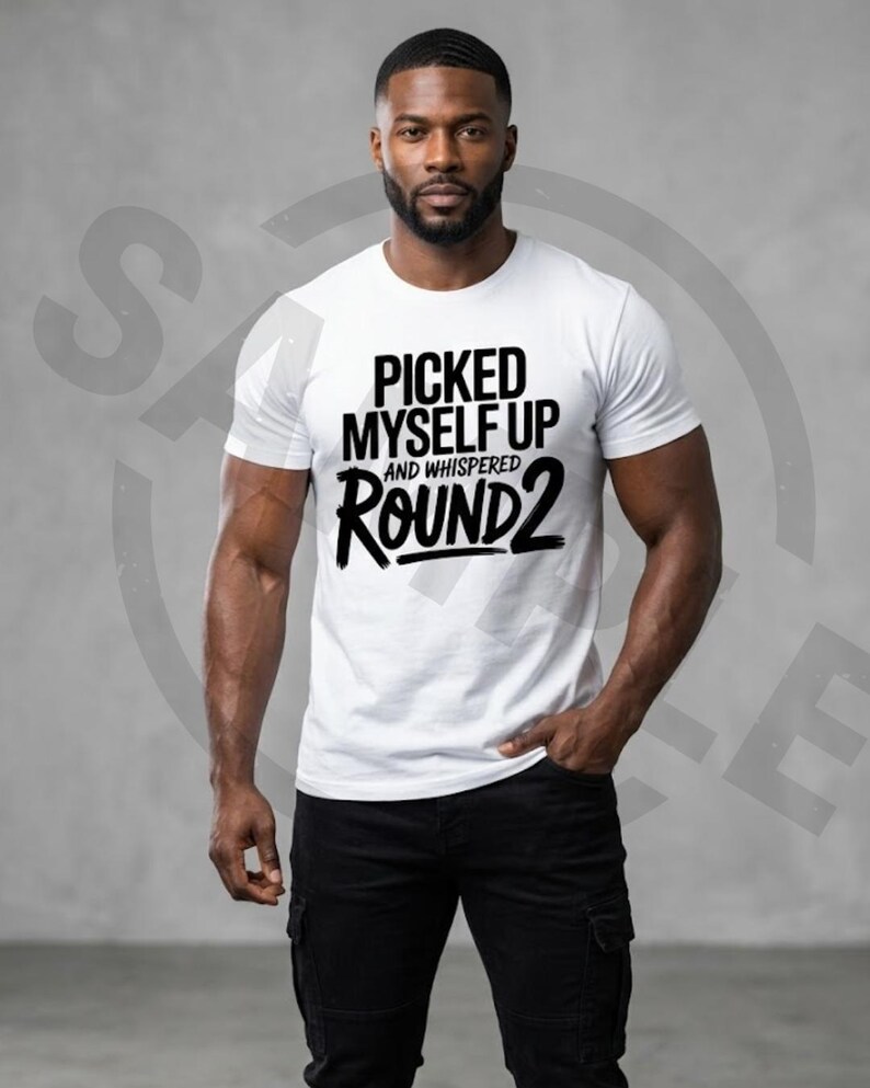 May include: A white t-shirt with the text "PICKED MYSELF UP AND WHISPERED ROUND 2" in black. The person is wearing black trousers.