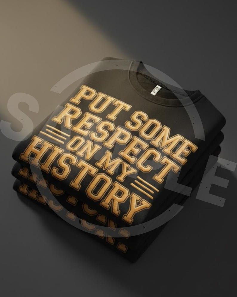 May include: A stack of black sweatshirts with the text "PUT SOME RESPECT ON MY HISTORY" in gold lettering. The sweatshirts are folded neatly, with the text prominently displayed on the top shirt.