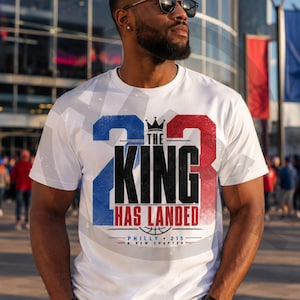 May include: A white short-sleeve t-shirt featuring a large graphic with the blue number 23 on the left and red number 23 on the right. The text reads THE KING HAS LANDED with a crown icon, a basketball graphic, and the text PHILLY 215 A NEW CHAPTER.