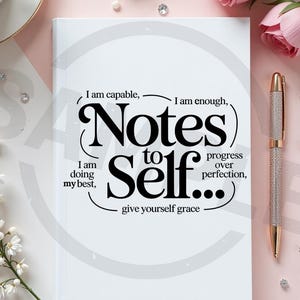 May include: A white journal with the text "Notes to Self" in a black, stylized font. The journal includes inspirational phrases like "I am capable" and "give yourself grace." A silver and gold pen rests on the right.
