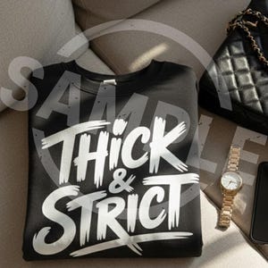 May include: Black sweatshirt with the words "Thick & Strict" in white, stylized lettering. The sweatshirt is folded on a light-colored surface, next to a black purse with a gold chain strap, a gold watch, and a black phone.
