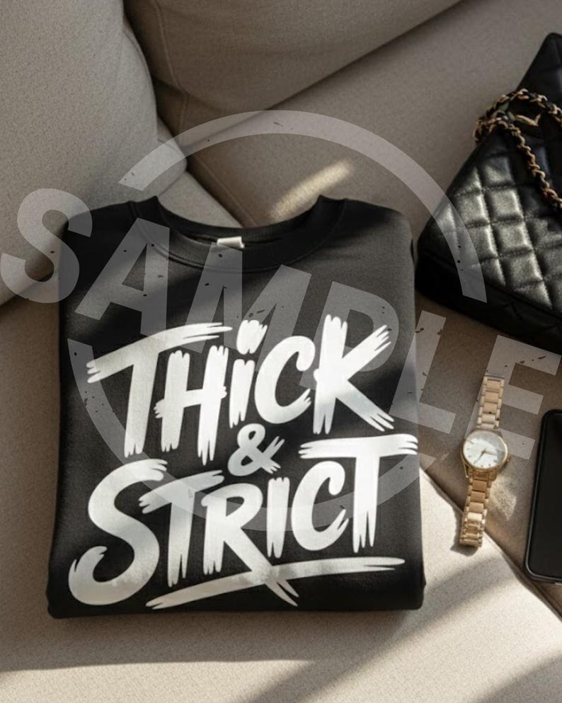 May include: Black sweatshirt with the words "Thick & Strict" in white, stylized lettering. The sweatshirt is folded on a light-colored surface, next to a black purse with a gold chain strap, a gold watch, and a black phone.