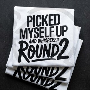 May include: Three white t-shirts stacked on a black surface. Each shirt features the phrase "PICKED MYSELF UP AND WHISPERED ROUND 2" in bold black lettering. The shirts are neatly folded.