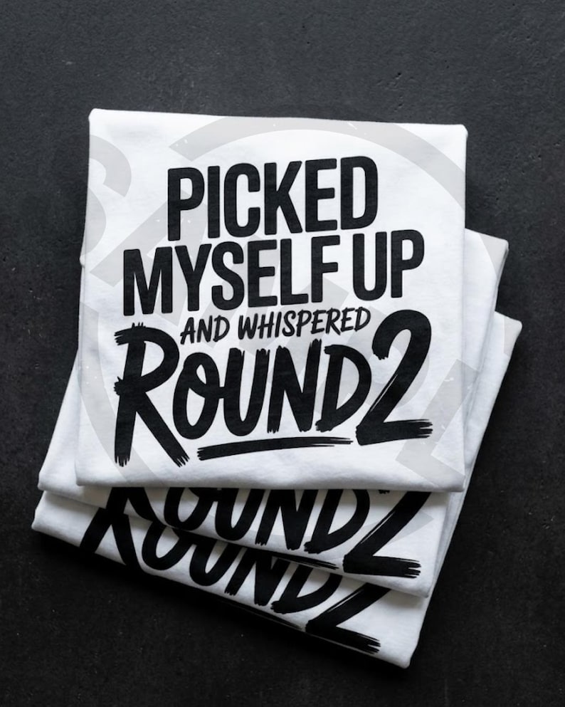 May include: Three white t-shirts stacked on a black surface. Each shirt features the phrase "PICKED MYSELF UP AND WHISPERED ROUND 2" in bold black lettering. The shirts are neatly folded.