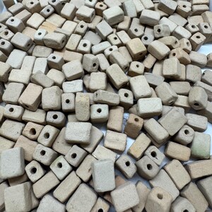 May include: A large collection of rectangular, light beige ceramic beads with a matte, textured finish. Each bead features a single, centered hole drilled through the middle, showcasing a uniform, earthy appearance and a consistent, blocky shape.