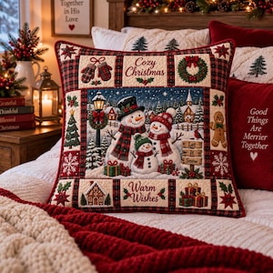 May include: A square decorative pillow featuring a central scene of three snowmen in a snowy landscape with a lamppost, church, and trees. The border includes panels with text reading Cozy Christmas, Warm Wishes, and Let It Snow, plus holiday motifs.
