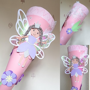Può includere: Un cono di carta rosa decorato con un design a fata. La fata ha una corona d'argento, un vestito viola e ali iridescenti. Un fiore viola e il nome "Emily" sono visibili anche sul cono. Il cono è probabilmente per la scuola o una festa.