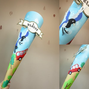 Può includere: Un cono di carta azzurra decorato con una gazza, un autobus rosso e uno striscione con la scritta "1. Klasse". Il cono è progettato per il primo giorno di scuola e presenta un design stravagante con alberi e nuvole.