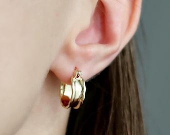 May include: A pair of gold hoop earrings. The earrings have a textured, organic design. The earrings are worn on an ear, with the focus on the jewelry. The earrings are gold in colour.