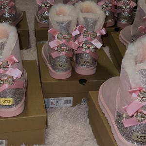 May include: Pink UGG boots with a sparkly silver design on the front and back. The boots have a bow detail and a fluffy white interior. Several pairs of boots are displayed on cardboard boxes.