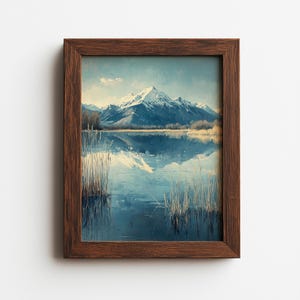May include: A framed landscape painting depicts a serene mountain scene. Snow-capped peaks are reflected in a calm lake, with reeds in the foreground. The artwork is encased in a dark brown wooden frame, creating a classic aesthetic.