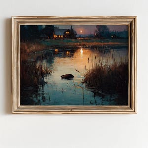 May include: A framed landscape painting shows a tranquil lake scene at dusk. The artwork has a golden frame. The water reflects the warm glow of a setting sun and the lights of a distant house. Reeds and grasses line the water's edge.