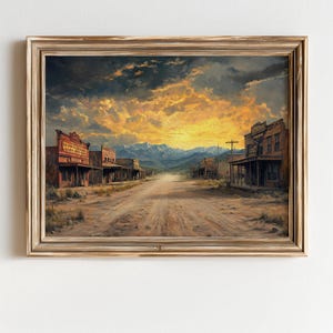 May include: A framed landscape painting of a Western town at sunset. The artwork depicts a dirt road leading towards mountains under a dramatic sky. Buildings line the road, and the overall color palette is warm with shades of brown, orange, and gold.