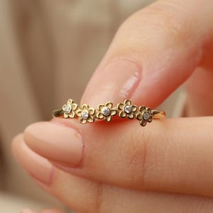 May include: A gold-colored ring featuring a floral design. Each flower has a small, clear gemstone in the center. The ring is held between fingers, showcasing the delicate details of the jewelry.