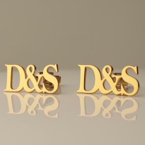 May include: A pair of gold-toned metal stud earrings featuring the letters D and S connected by an ampersand symbol. The polished surface of the letters reflects light, creating a bright appearance for these custom initial jewelry pieces.