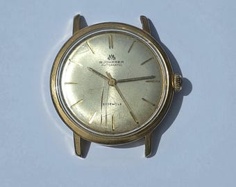 Puede incluir: Un reloj de pulsera Bucherer Automatic en tono dorado con esfera redonda color champán, marcadores de hora y manecillas doradas. La esfera muestra el texto Bucherer Automatic y 21 Jewels. La caja tiene cuatro asas y una corona lateral.
