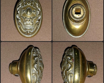 Puede incluir: Un pomo de puerta ovalado de latón con un intrincado patrón de volutas en relieve en la cara frontal. La parte posterior muestra un orificio cuadrado central para el eje de montaje. El perfil lateral muestra un borde redondeado.
