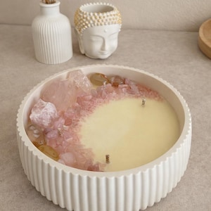 Peut inclure: Un bougeoir blanc à bordure cannelée rempli de cire jaune crème et orné de cristaux roses. La bougie a trois mèches et est entourée de cristaux décoratifs. Une tête de Bouddha et un vase blancs sont en arrière-plan.