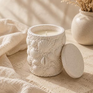 Könnte beinhalten: Eine weiße Kerze in einem strukturierten Behälter, der mit Muscheln verziert ist, mit passendem Deckel. Die Kerze steht auf einer Steinoberfläche, neben einer Vase mit getrockneten Blumen. Das Thema ist maritim und neutral.