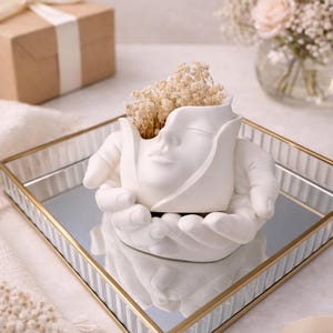 Puede incluir: Una maceta de cerámica blanca con forma de rostro sostenida por manos, llena de flores secas, sobre una bandeja espejada con borde dorado. Una caja de regalo con una cinta y un jarrón de flores en el fondo.