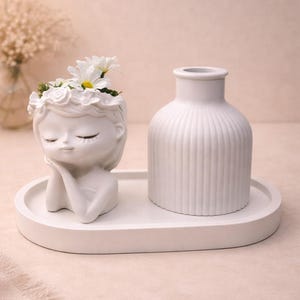 Könnte beinhalten: Ein weißer Keramik-Blumentopf in Form eines Frauengesichts mit Blumenkranz, der das Kinn hält. Daneben eine weiße, geriffelte Vase. Beide stehen auf einem weißen ovalen Tablett. Weiße Blumen befinden sich im Pflanzgefäß.