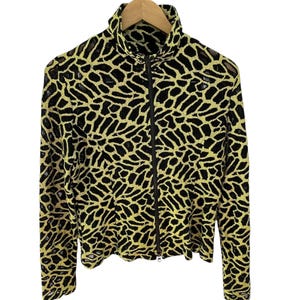 May include: A black and gold zip-up jacket with a high collar. The jacket features a repeating pattern of black shapes on a gold background. The jacket is hanging on a wooden hanger against a white background.