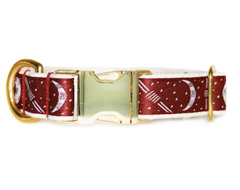 May include: A pet collar featuring a deep red fabric strap with a white pattern of crescent moons and small dots. The collar includes a shiny gold-toned metal side-release buckle, a matching gold D-ring, and a gold-toned metal adjustment slider.
