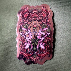 May include: A sticker with a detailed floral design in shades of pink, red, and green. The sticker has a brown border and is set against a green background. The design features symmetrical floral elements.