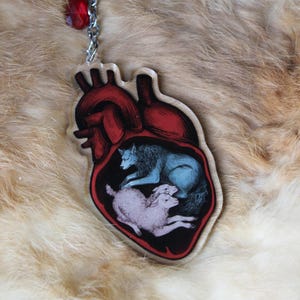 May include: A heart-shaped keychain with a red and black anatomical design. Inside the heart, a blue wolf and two pink sheep are depicted. The keychain has a silver chain and a red crystal accent.