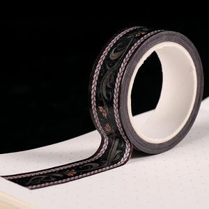 May include: A roll of decorative washi tape with a black background and a floral design in shades of brown and gray. The tape is unrolled on a white dotted paper.