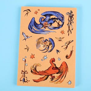 Puede incluir: Una hoja de pegatinas con ilustraciones de aves míticas en tonos azules, naranjas y dorados. Las pegatinas incluyen varios diseños de aves, estrellas y elementos decorativos sobre un fondo beige.