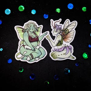 May include: Two sticker illustrations of fairies with green skin, wings, and floral accents. One fairy wears a red top and the other a green top. The background is black with scattered blue and green sequins.