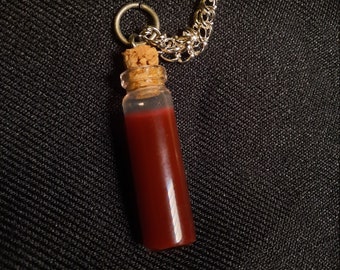 May include: A small glass vial filled with a deep red liquid, sealed with a cork stopper. The vial is suspended from a silver-colored chain necklace. The background is a dark, textured fabric.