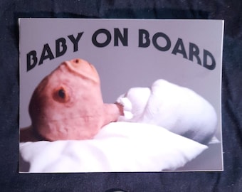 May include: A sticker with the text "BABY ON BOARD" in black letters on a gray background. The sticker features a photo of a baby lying on a white blanket.