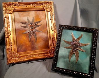 May include: Two framed artworks. One has a gold ornate frame, the other a black frame. Both feature a central starburst design with metallic wings and jewel accents. The background colors are a mix of orange, brown, and teal.