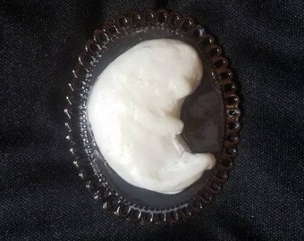 May include: A white crescent moon shaped resin brooch with a black background, set in a black metal frame with a decorative border.
