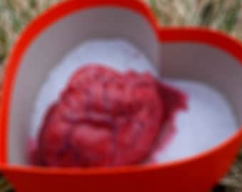 May include: A red heart-shaped box with a red, fleshy object inside. The box is sitting on a bed of dry grass.