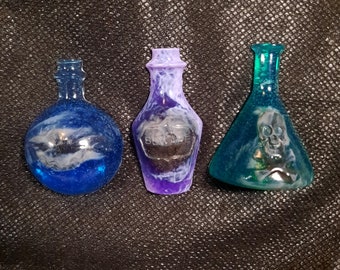 May include: Three glass bottles with a potion theme. The bottles are filled with a blue, purple, and green liquid. The bottles have a skull, a swirl, and a label that says "Beware" on them.