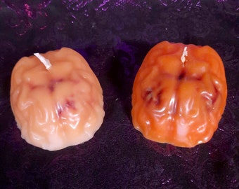 May include: Two brain-shaped candles, one orange and one light peach, on a purple background. The candles have a realistic brain texture and a small wick on top.