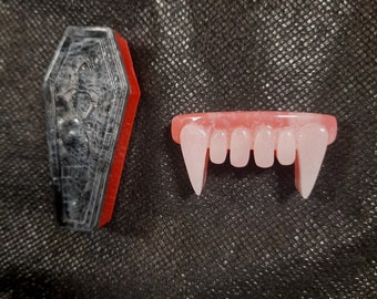 May include: A pair of plastic vampire fangs, with a pink base and white fangs, next to a small black and gray coffin-shaped box with a red border.