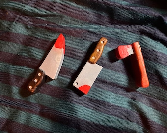 May include: Three toy knives with red painted blood on the blades. The knives are a butcher knife, a kitchen knife, and a small axe.