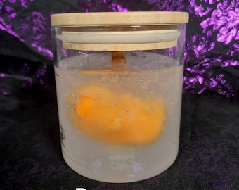 May include: A clear glass jar with a wooden lid containing a yellow beeswax block. The jar is sitting on a purple fabric background.