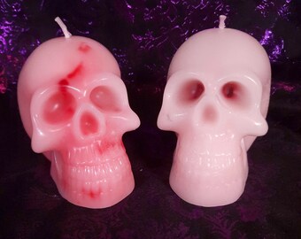 May include: Two pink skull shaped candles. One candle has a red streak down the side of the skull.