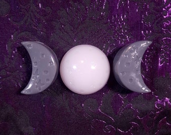 May include: A decorative set featuring a white sphere flanked by two gray crescent moons. The moons have star and dot details. The objects are arranged on a purple patterned fabric.